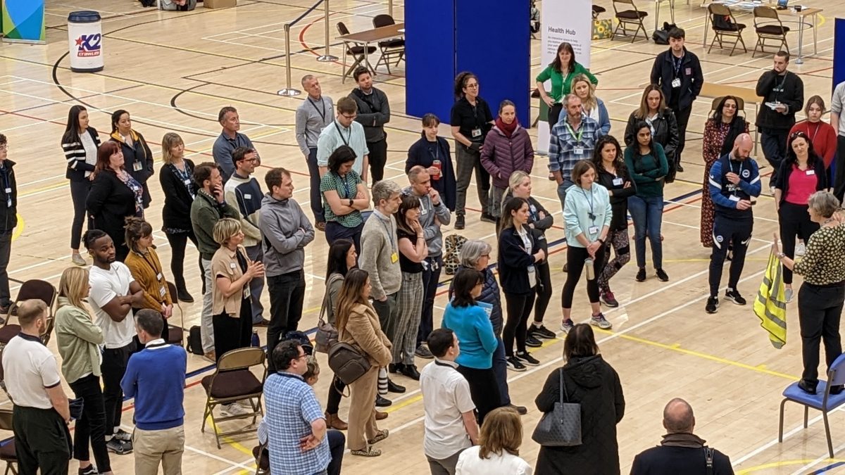 A large number of staff stood in a sports hall at a Community Appointment Day listening to someone speaking