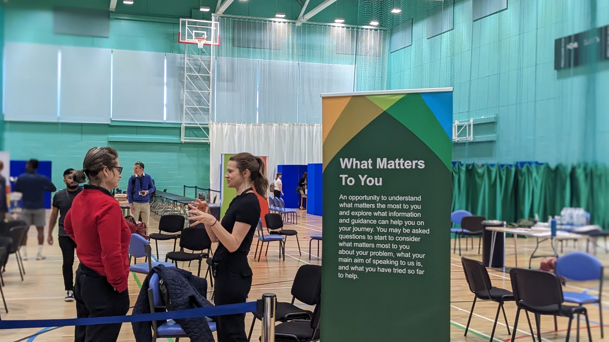 Photo of a sports hall with a woman stood helping to direct people where they need to go, next to a banner that reads 'What Matters To You'