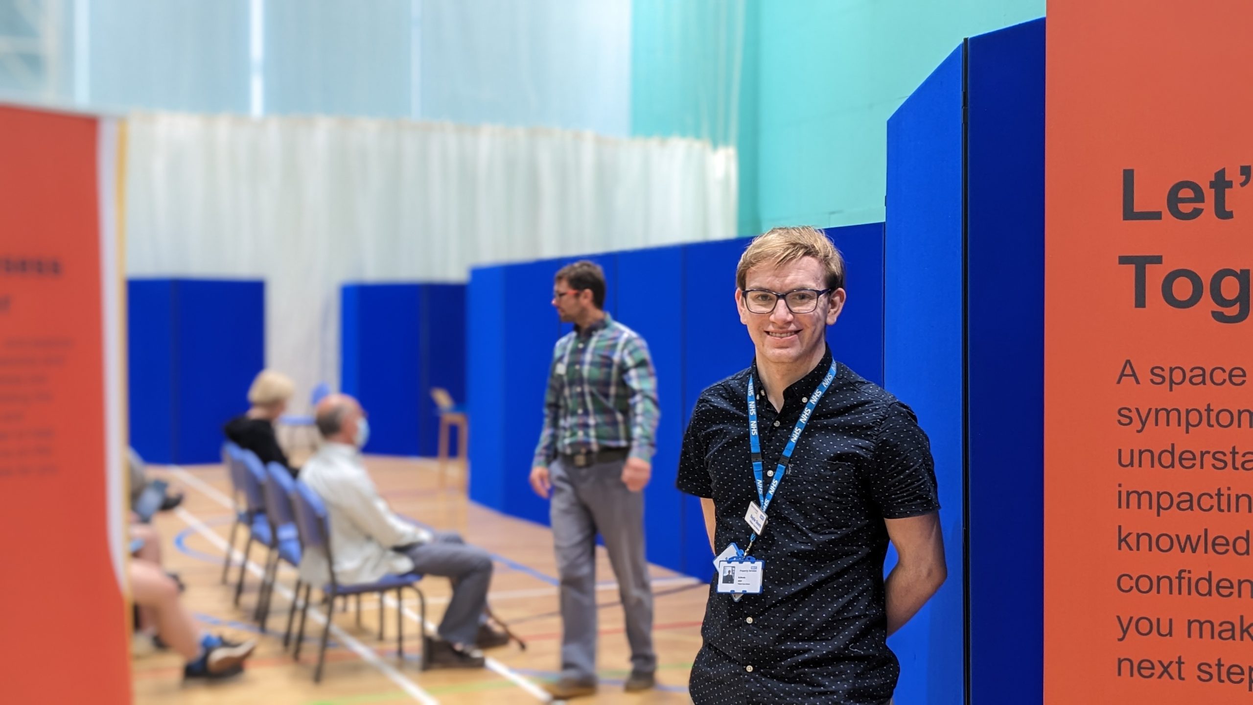 A younger white man wearing glasses, stood with his arms folded behind his back and smiling - he's stood in a sports hall at a Community Appointment Day