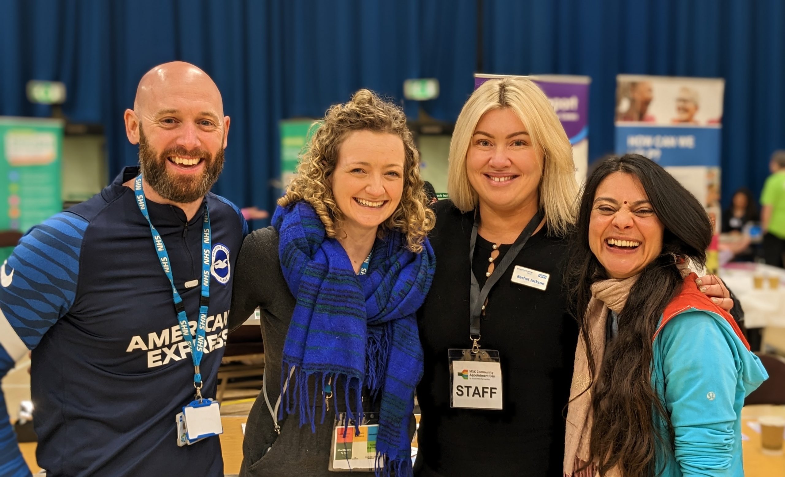 A group of four team members, one male, three female stand close together and smiling in a sports hall at a Community Appointment Day