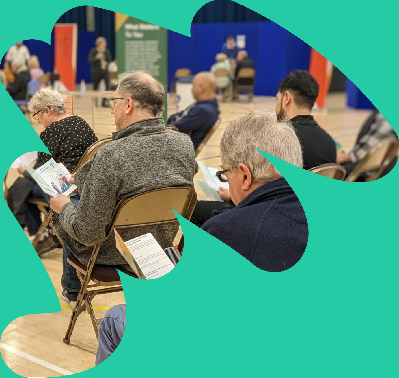 Image features people sitting on chairs in a sports hall reading information about the community appointment day they are attending.