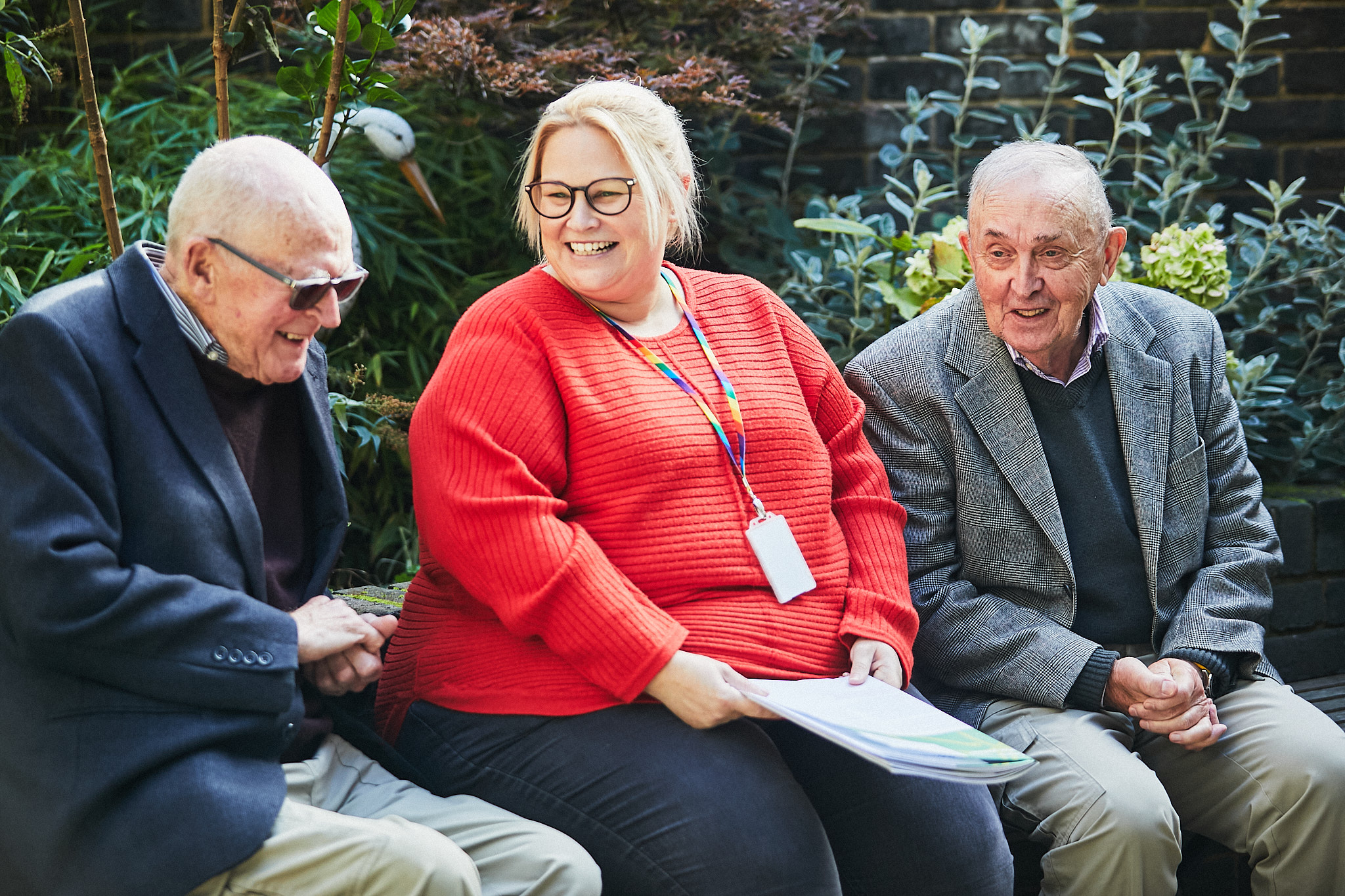 Sam Stevens, Memory Assessment Service Manager, a white woman wearing glasses sat outside with two Memory Assessment Service service users on her left and right - two older men, one wearing glasses and one without