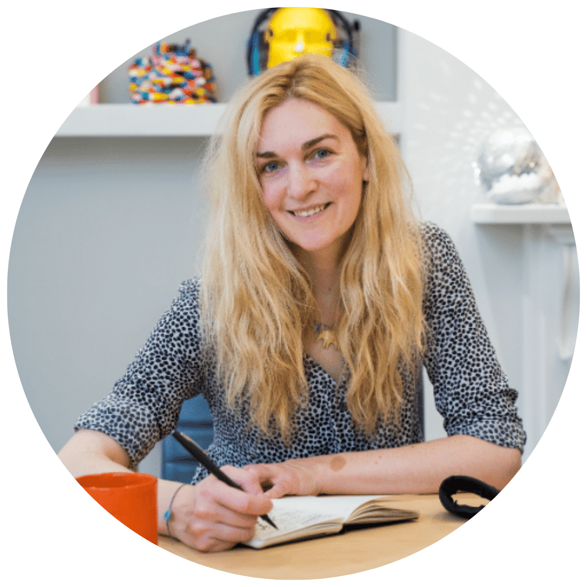 Photo of a woman with long, light coloured hair, smiling whilst sat at a desk holding a pen