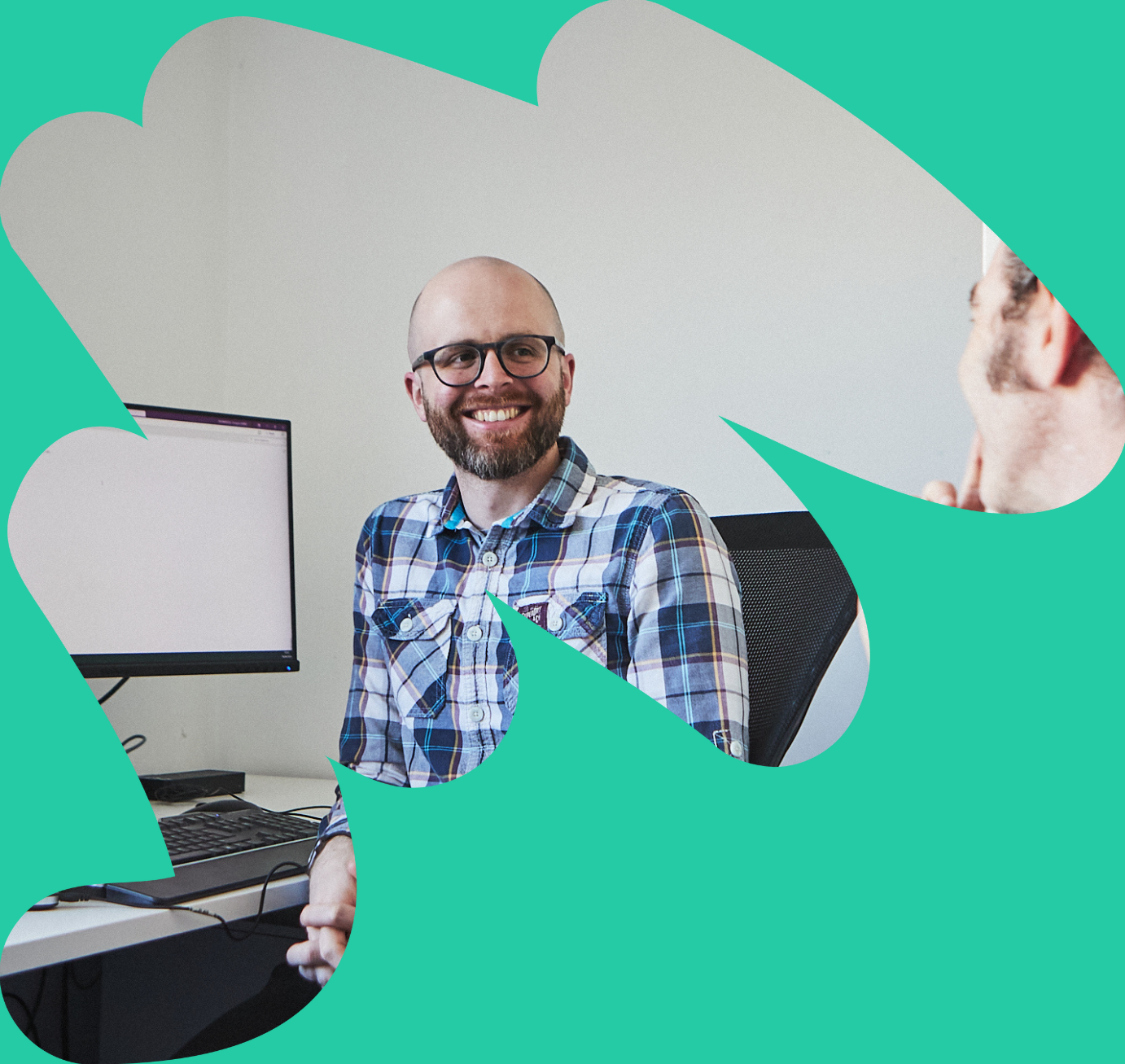 One of the Data and Analytics team - a white man with a beard and glasses, sat in front of a computer and smiling at another team member out of shot