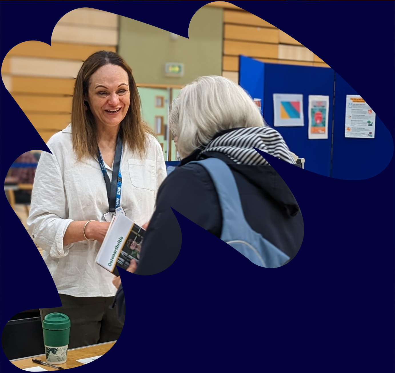 Image of an older woman at a reception desk checking in to a community appointment day and talking to a white female receptionist wearing a lanyard