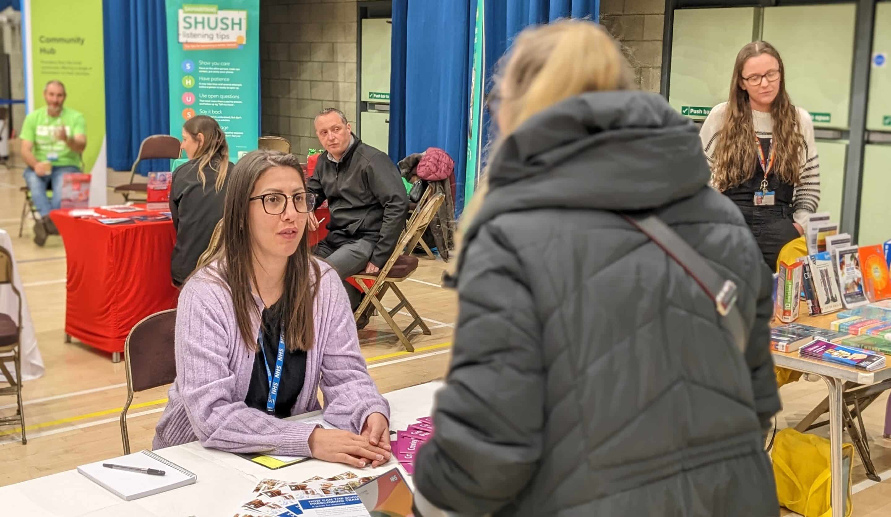 A woman wearing a dark coloured coat is stood at a desk talking to another woman wearing glasses who is sat down. They are in a sports hall at a Community Appointment Day