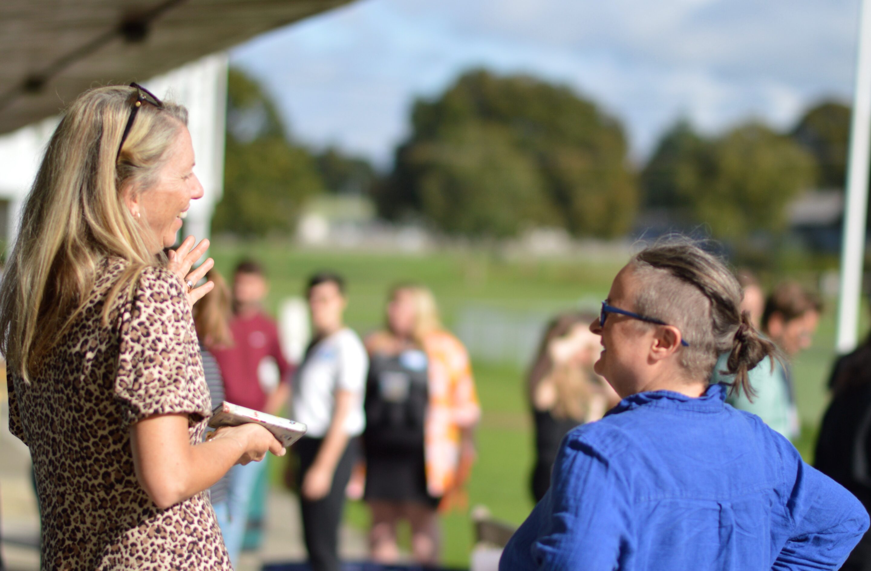 One woman with long blonde hair is laughing with another woman with dark hair tied up and with the sides of her head shaved wearing glasses