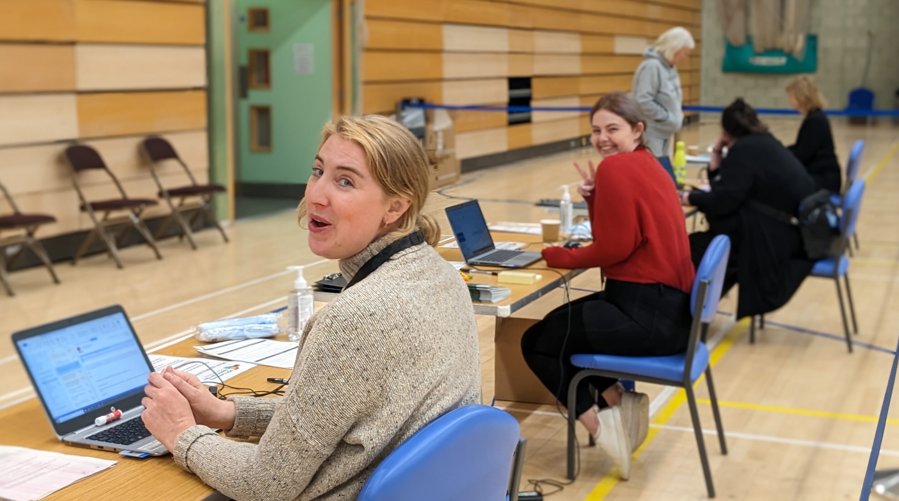 Two people sat at desks in a sports hall at laptops looking over their shoulders and smiling