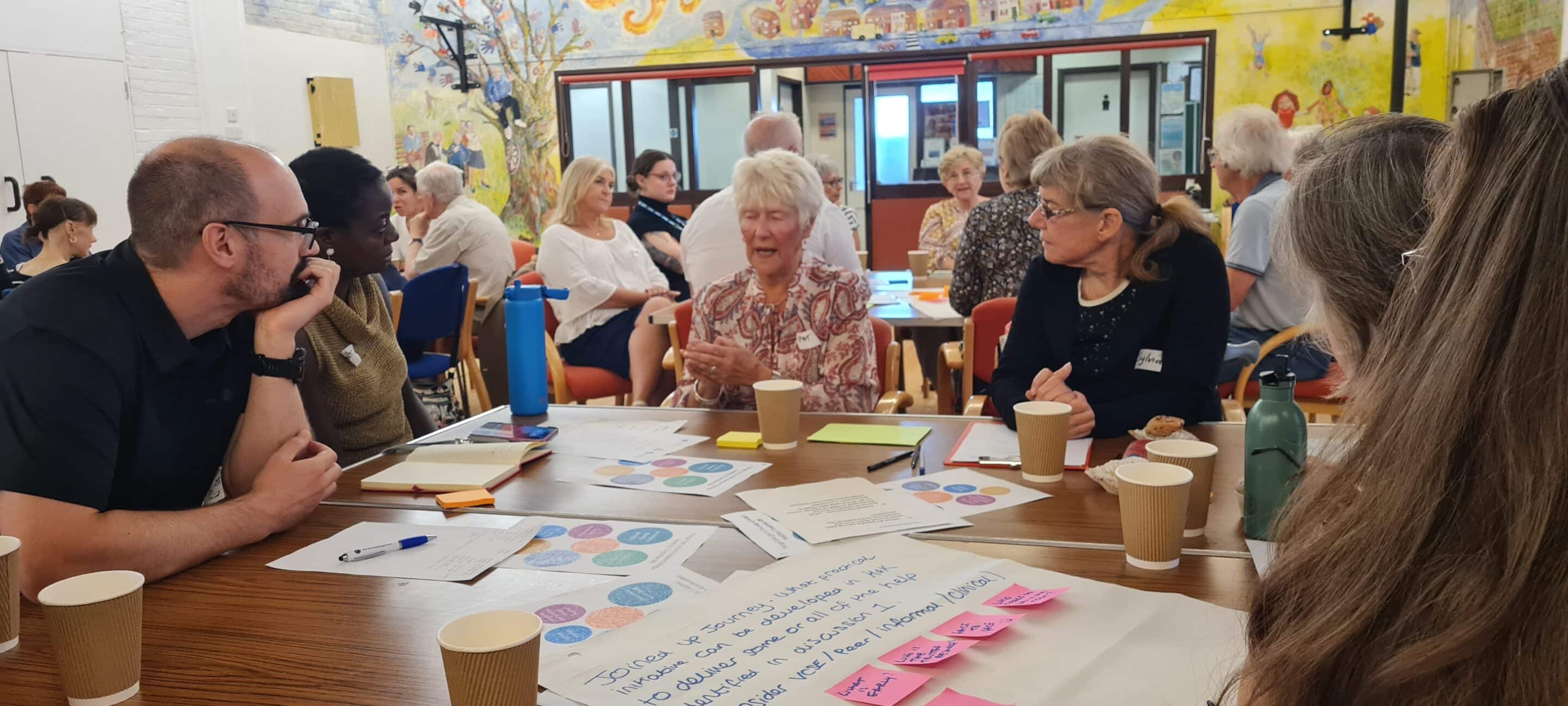 20250730_113820 (1) A group of people of various ages sit around a table in a bright, community-style room, engaged in lively discussion. Papers, pens, coffee cups, and post-it notes are spread out on the table. A colourful mural and more participants are visible in the background, contributing to a collaborative and inclusive atmosphere
