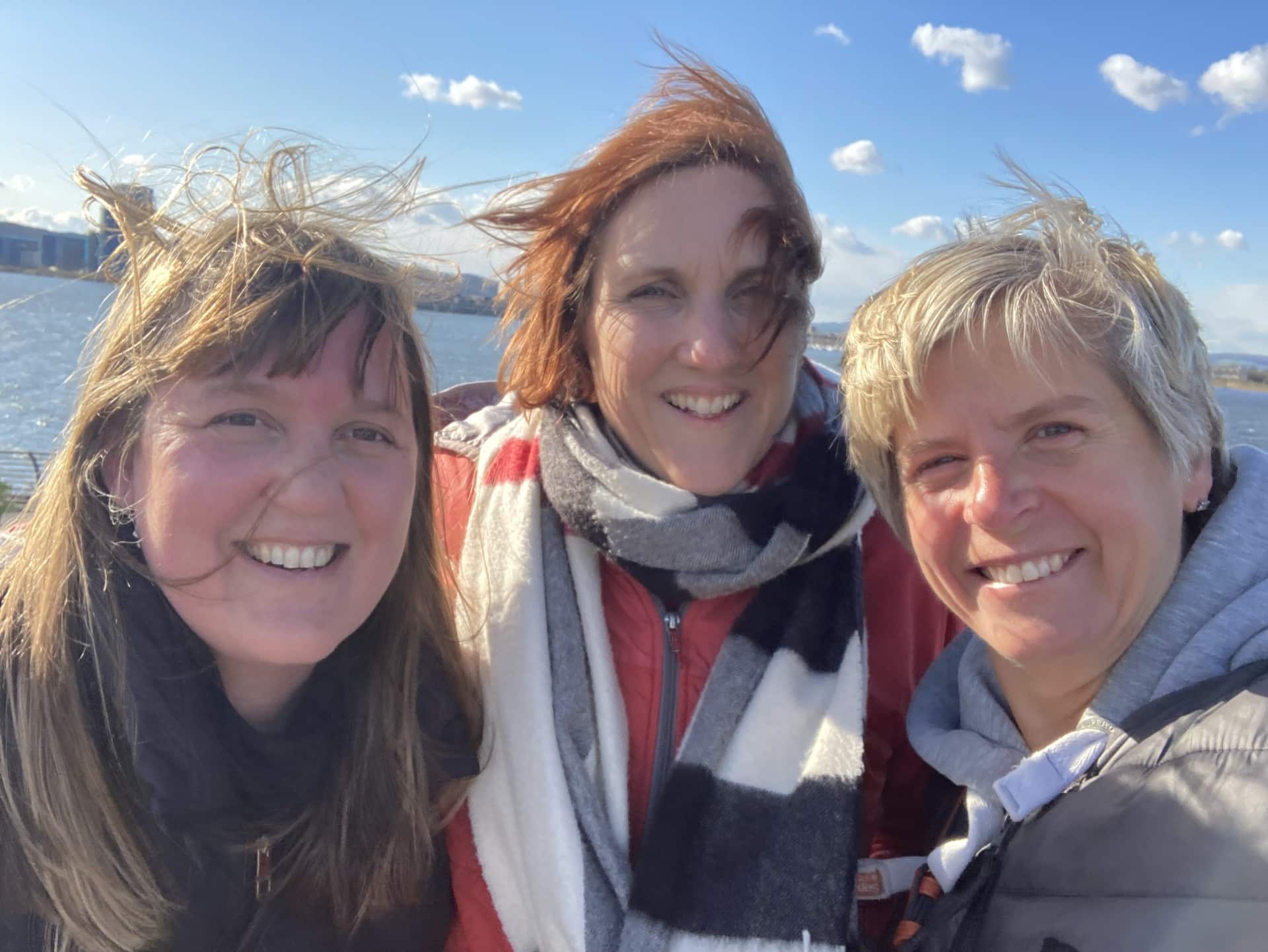 Photo of two white women outside with trees in the background. stood next to each other and smiling towards the camera. The woman on the left has long hair and the woman on the right has a short bobbed hair cut.