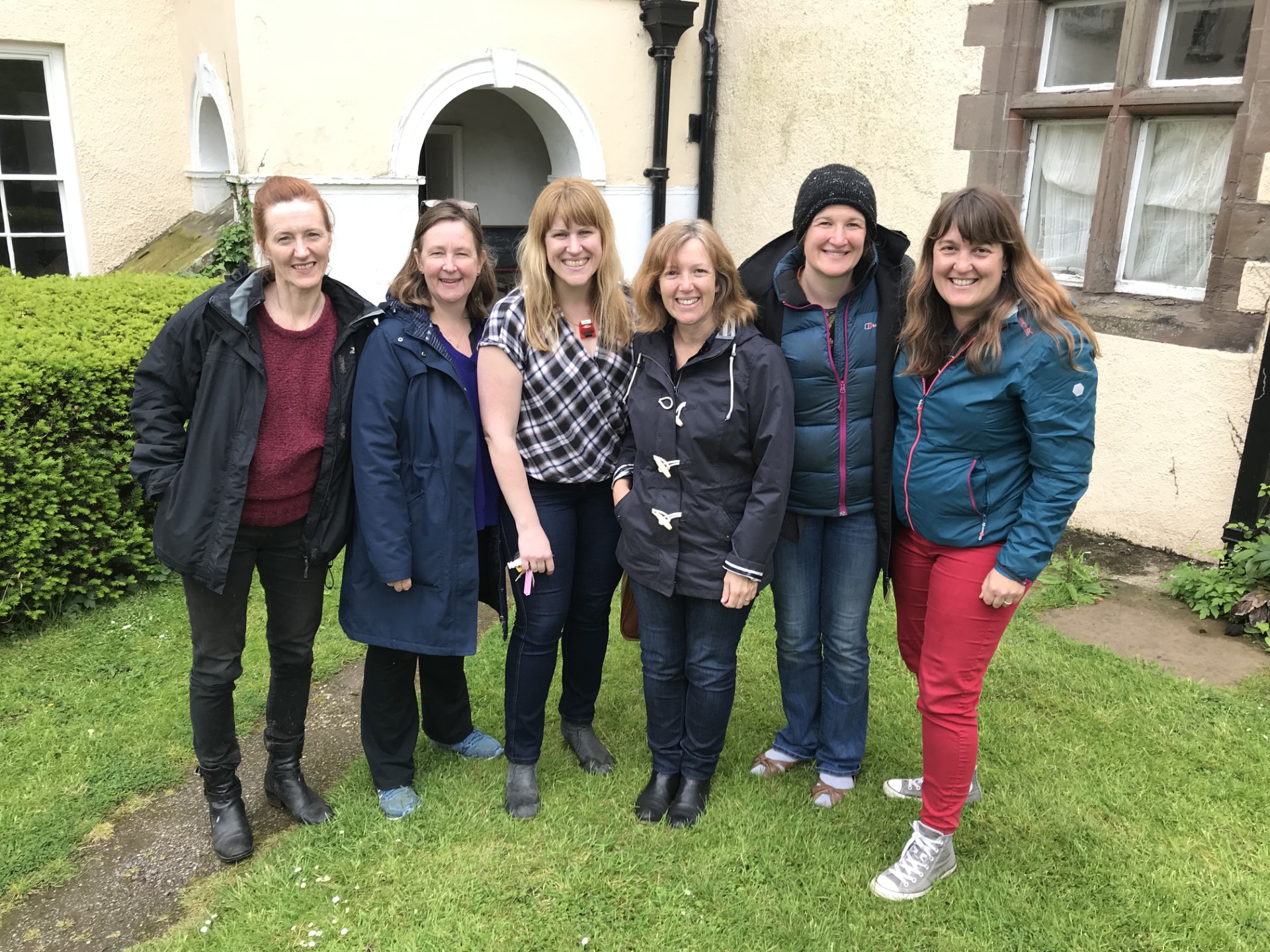 Photo of two white women outside with trees in the background. stood next to each other and smiling towards the camera. The woman on the left has long hair and the woman on the right has a short bobbed hair cut.